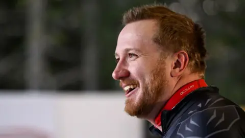 A man wearing a black, form‑fitting athletic suit with geometric panels stands outdoors in a snowy setting. The man faces sideways, looking toward someone off‑camera, with blurred winter trees and a partial event banner in the background.