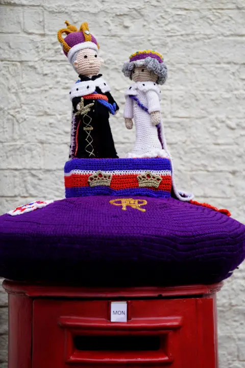 PA Media A woollen post box topper sees figures of King Charles and Queen Camilla standing on a plinth on top on top of a purple post box covering