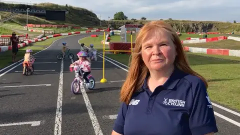 Coach Fiona Graham standing on the kart racing track, with small children practicing their cycle skills behind her. She is wearing a blue polo shirt with British Cycling on it, and has ginger hair reaching below her shoulders.