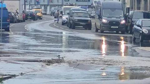 A road with water gushing up through a gap, and queues of traffic