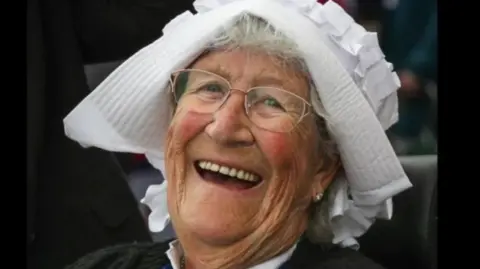 A smiling woman wearing glasses and a white headpiece that resembles a bonnet. Grey hair is visible beneath  the head covering and she has a dark top on. 