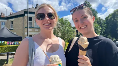 Two women smiling into the camera. The woman on the right has blond hair and sunglasses on. The woman on the right has brunette hair with sunglasses on her head. They are both holding ice cream. They are standing in front of a leafy avenue, with houses and a black marquee visible.