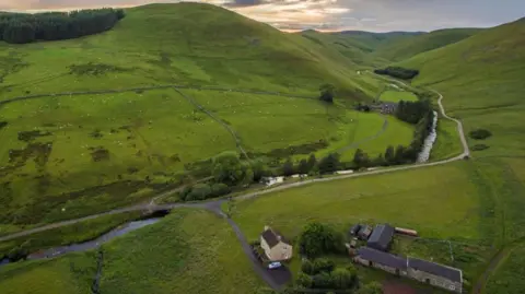 An aerial view of a farm in Upper Coquetdale with a road running alongside the river Coquet. The farm with associated farm buildings is set back off the road. Beyond are lush green hills and the hint of the sun behind the clouds.