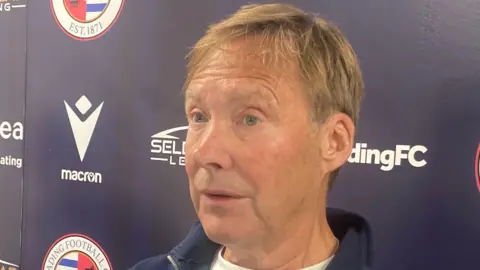 A picture of Nigel Howe sat in front of a Reading FC banner and wearing a blue jumper and white T-shirt. He has short, brown hair.