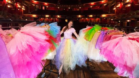 Katie Sparkes wearing a tutu on stage at the Bristol Hippodrome, surrounded by clothing rails of other tutus