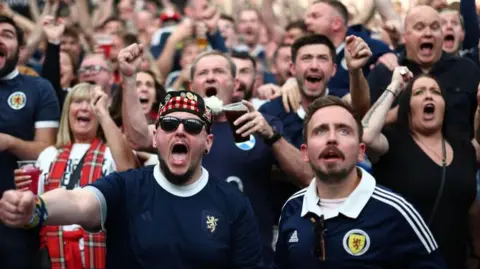 Large crowd of football supporters wearing dark blue Scotland tops, gathered closely together in an indoor venue, many holding plastic cups of beer with arms raised, including a person in the foreground wearing a tartan hat.