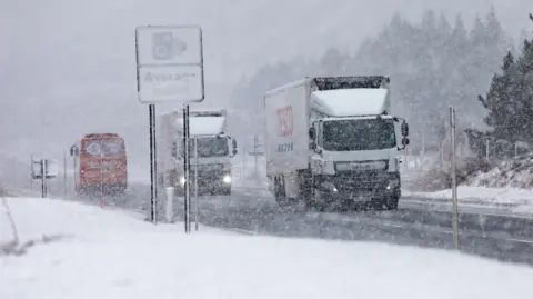 Lorries make their way along a snowy A9 with a sign for average speed cameras only just visible