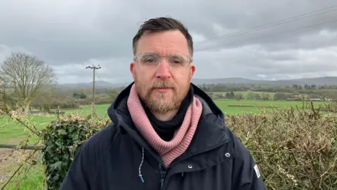 A man with glasses, a pink scarf and a black jacket with fields in the background on a cloudy day.