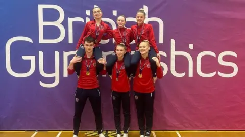 Three girls are sitting on the shoulders of one boy and two girls. They are wearing red tops and medals. Behind them is a large sign that says "British Gymnastics" on it.