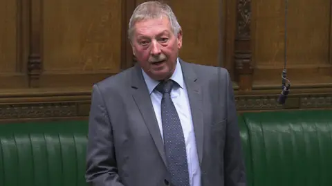 Houses of Parliament Sammy Wilson speaking in the House of Commons. He has a grey suit on with a blue polka dot tie and blue shirt. He has gray balding hair and a gray moustache. Behind him are green leather seats.