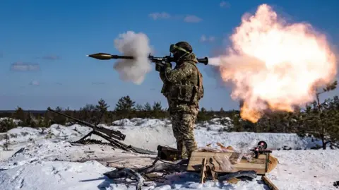 Getty pictures of a soldier faces the left and fired a weapon in the same direction, and fired a ball from the fire behind it. The land is icy and there is a gun next to the soldier. The sky is blue.