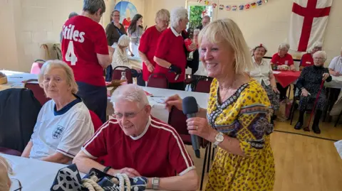Helen Taylor/Yaxley Parish Council Vernon, a man with white hair, wearing a red football shirt, sitting on a table. A woman in a yellow dress who is holding a microphone is standing next to him, chatting to him.