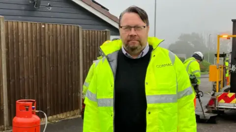 The image shows a bearded man in a hi-vis jacket looking at the camera. He is standing outside. There is a fence behind him to the left. To the right, there is a workman with a tool and machinery on the road.