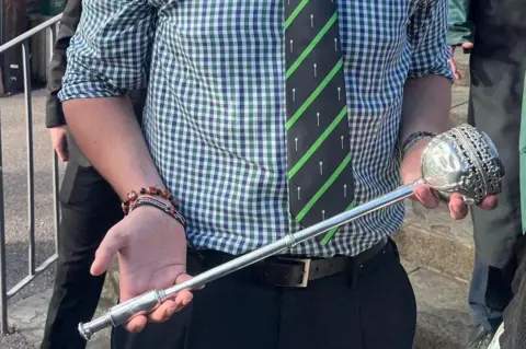 Caleb Tutt A silver mace being held by a man. All you can see is the mace and the hands of the man, as well as his torso. He is wearing a checkered white, purple and green top with a striped dark green and light green tie.