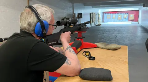 Tom Jackson/BBC A man is wearing blue headphones while holding a rifle. He is aiming for a target at the other end of a long brick walled room.