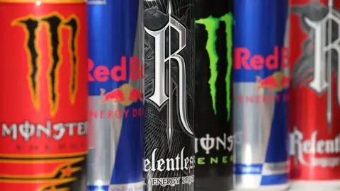 Close up image shows brightly coloured Monster, Red Bull and Relentless energy drinks are displayed on a supermarket shelf. 