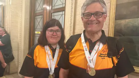 Claire O'Neill and Eamonn Brummitt smile at the camera, both wearing the yellow and black jerseys of the Special Olympics Ulster team and each wearing two medals around their neck. Claire has shoulder-length dark brown hair, while Eamonn has short grey hair and is wearing glasses.