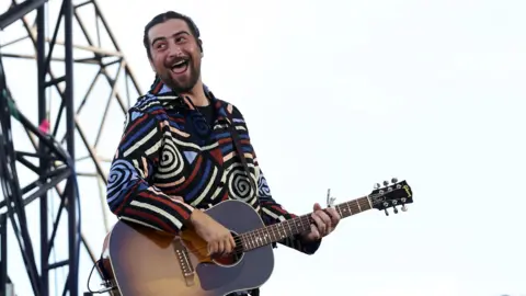 EPA A man looks over his shoulder smiling as he sings. He is playing an acoustic guitar. He's wearing a bright, long-sleeved top with swirling colourful patterns on it.