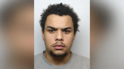 West Yorkshire Police A man aged around 20 with frizzy hair and furrowed brows. He is sporting an cross expression on his face while staring into the camera.