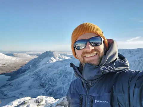 Matt Cooper A man in a grey/blue jacket with a yellow beanie and sunglasses on smiles for a selfie in front of a snow-topped mountain