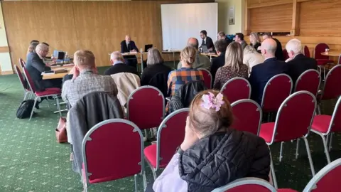 Andrew Turner/BBC John Dowsett, the planning inspector sits at his desk at the front of the inquiry room, with severa people seated on red steel-framed chairs. The back wall is timber panelling and the carpet is green. Counsel for Breedon Trading is on the left of the image, and Norfolk County Council on the right.