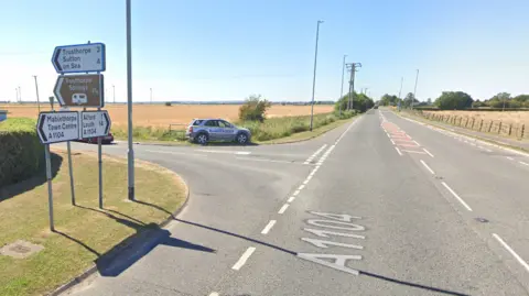 A Google Street View image capture of the A1104 at the junction of the A1031. There are direction signs and a car with advertising on parked at the junction. It is a rural location with fields stretching out into the distance.