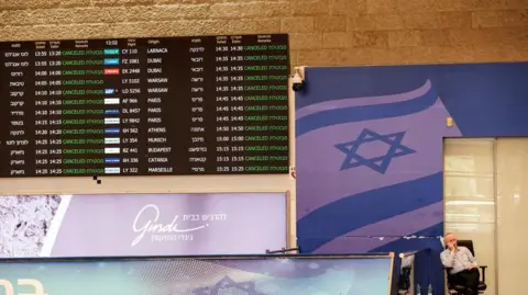 A staff member sits next to a departure board where a number of flights listed are shown as cancelled.