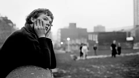 A black-and-white photo of a boy leaning on a wall with his hands framing his face. He is looking to the right and is wearing a dark jumper. The background is out of focus but shows a group of about five people looking in the direction of the camera. They are standing on a patch of grass in the middle of a housing estate.