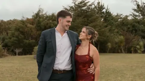 Cai James Cai and Annie smiling looking at each other stood in a field with greenery behind them. Cai, on the left, wears a white shirt and navy blazer. Annie, on the right, wears a red dress and has short brown hair.