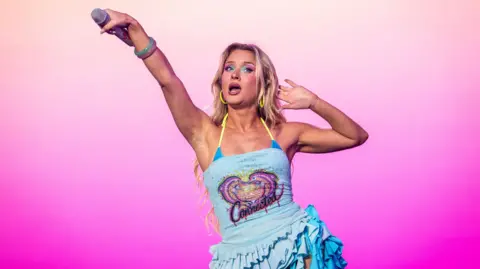 BBC Zara Larsson performs on stage and she is holding a microphone outstretch to the crowd. She wears a blue sparkly dress.