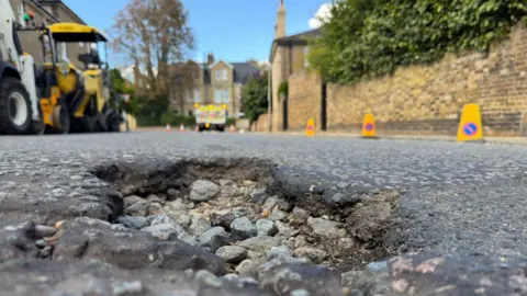The photo is taken from the road level in a residential street. It shows a pothole in the foreground, around 8cm deep, where the asphalt surface has broken away, leaving course rubble below. In the background, machinery and highways vehicles have yellow lights flashing as they prepare to repair it.