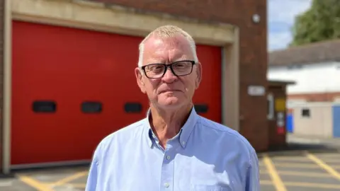 Man in his 60s, in a pale blue shirt and dark rimmed glassed. He is standing in front of a red fire station garage door, lit by the sun. 