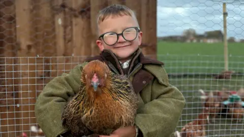 A seven-year-old boy smiling at the camera while holding a chicken.