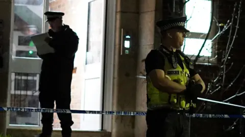 Police officers stand outside a flat. One is guarding a cordon, one is looking at a clipboard and two others are talking.