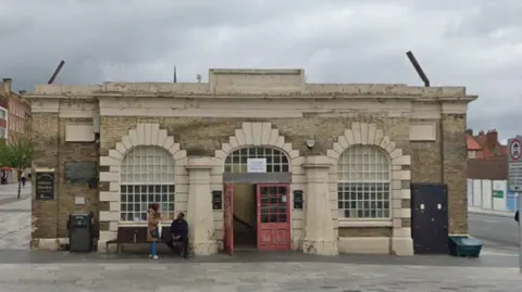 An outside view of the Stockton Shambles building. It has open red doors in the centre of stone archways with windows in them. Two people are near the bench to the left of the front doors.