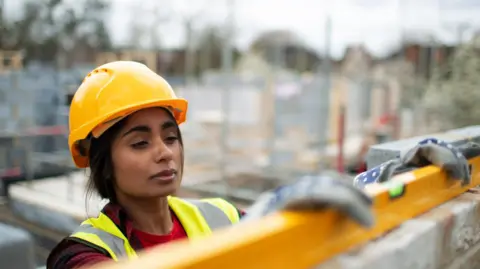 South Asian woman in hard hat and hi viz uses a spirit level on a brick wall. She is outside on a building site.