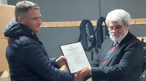 York City Rowing Club A man in a navy coat receiving a certificate from a man in a black suit with a red and blue tie.