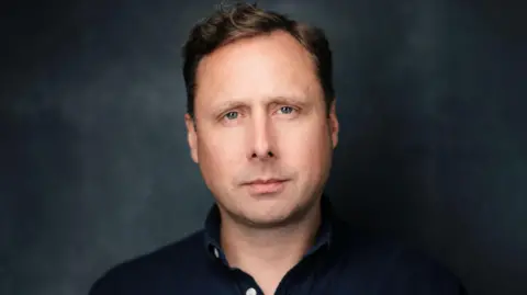 A headshot of Adam Graham, who is wearing a navy shirt. He has short brown hair and is looking at the camera without much expression. Behind him is a blurry grey and black background.