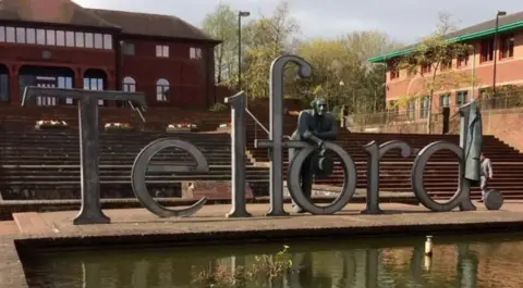 BBC A large metal sculpture which spells out the word Telford with a metal sculpture of a man leaning on it, a small pond in the foreground and a number of large buildings at the top of a flight of steps