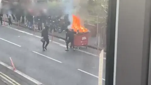 Tejal Samat and Nishath Mohammed Picture take from window showing two people in dark clothing setting fire to a bin. A crowd of people are standing on the footpath.
