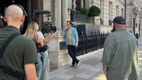 Quentin Tarantino is seen wearing a blue jacket and white t shirt, leaning against the wall of a hotel. In front of him is someone with a camera, while support crew are also standing around