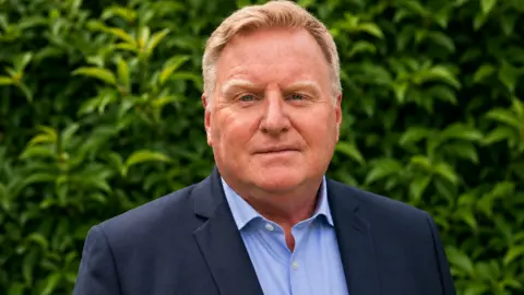 Andy Woolley has short blond hair and wears a navy blue suit and a light blue shirt.
