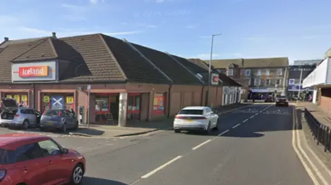 Google Fowlds St in Kilmarnock, shows a red and white car driving down the street an Iceland supermarket is to the left and a car park is on the right.