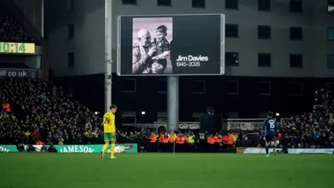 A black and white image of Jim Davies with a scarf round his neck holds a young boy in a football kit in his arms appears on a big screen above the pitch. To the left of the photo is a timer that says 80:34. Two players are on the pitch and match officials in orange and yellow high vis jackets stand underneath the screen and the stands are full.