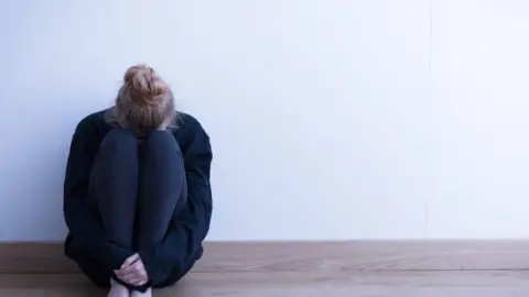 Getty Images A woman sits curled up on the floor, her head between her knees