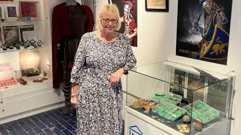 Lorraine is wearing a blue and white paisley dress and is leaning on a display cabinet that has some of her finds inside. She is wearing glasses and is smiling at the camera.