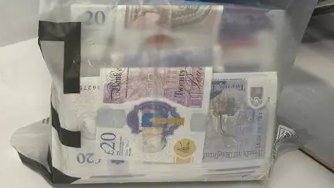 A wodge of £20 notes in a clear plastic bag is held up for the picture.