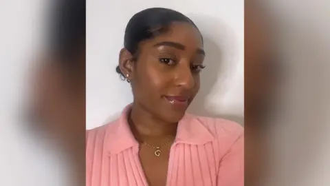 Grace Henry A woman poses for a selfie against a white backdrop. Her hair is tied back, head turned slightly to the side. She wears a ribbed salmon pink top with a collar, open at the neck to reveal a short gold necklace with a capital G pendant.