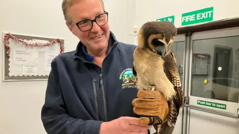 The photo shows handler, Tim Sheppard, standing indoors wearing a brown leather glove. On his hand is a brown owl. Tim is smiling.