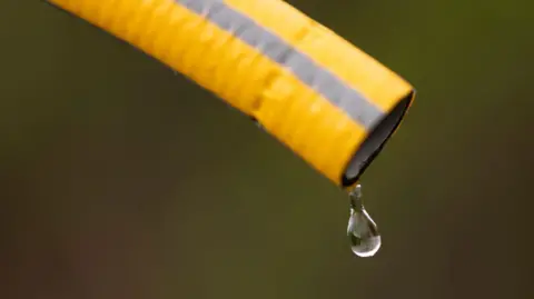 A yellow hosepipe with a grey stripe running down it and one water drop appearing out the end